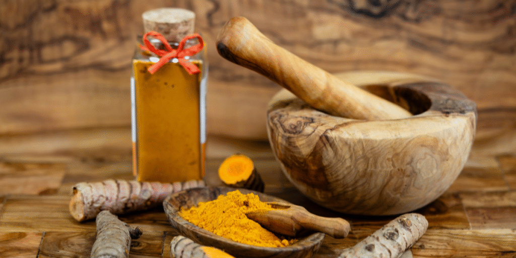 Wooden bowl and bottle filled with organic turmeric powder and fresh turmeric roots on a wooden table, with leafy background